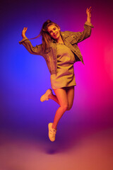 Fototapeta na wymiar Portrait of young girl in casual cloth jumping, posing isolated over gradient pink blue background in neon light. Cheerful look
