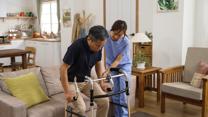 asia female nursing aide helping injured retired male to use a walker while he is slowly standing...