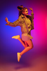 Portrait of young girl in casual cloth jumping, posing isolated over gradient pink blue background in neon light. Happiness and joy.