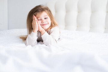 beautiful happy little kid girl with blonde hair having fun lying on white bed linen