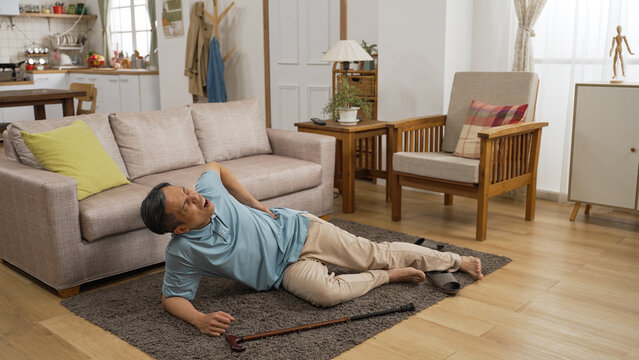 old Asian grandfather as he is sitting on floor groaning in pain after fall in a living room at home.