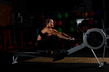 Serious young lady exercising on rowing machine in dark gym