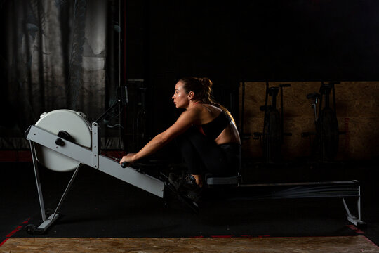 Serious young lady exercising on rowing machine in dark gym