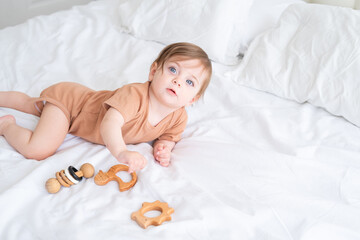 baby girl with blue eyes in brown bodysuit playing with wooden toys on white bedding on bed