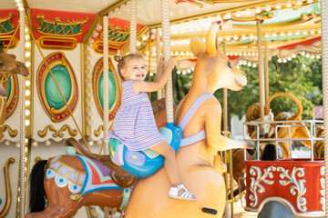 funny little kid girl in colorful dress rides on carousel in an amusement park in summer day