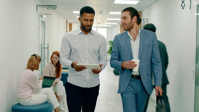 Two important looking men with beards pass people through the large office while talking about business stuff, they are both wearing suits and look very intelligent