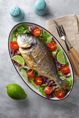 Baked whole dorado fish on a plate with fresh vegetables on the light gray background top view vertical photo