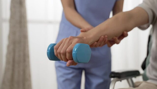 slow motion with closeup hand of wheelchair user holding lifting dumbbell with nurse’s support at background. home health care and fitness for the disabled concept