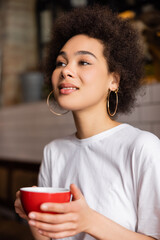 cheerful african american woman in hoop earrings holding cup of coffee