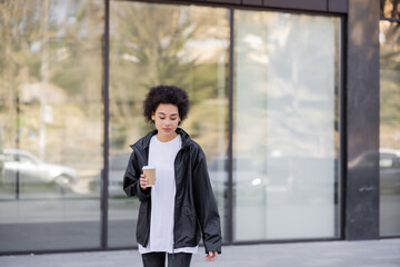 curly african american woman in jacket holding paper cup on urban street