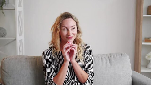 Evil plan. Ironic woman. Sarcastic expression. Pretty smiling lady running fingers full of malicious joy sitting sofa light room interior.