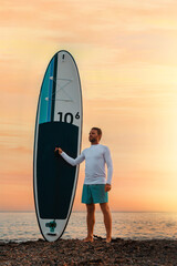 Extreme activity. Fit man holding a sup board and posing on the beach. In the background, the ocean and the sunset. Vertical. Copy space. Summer surfing