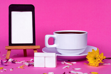 Chalkboard with white coffee cup, wooden block and flowers on pink background.