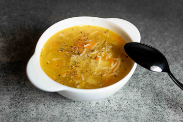 Chicken soup with noodles, potatoes, onions and carrots in a white deep plate on a gray background.