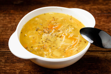Chicken soup with noodles, potatoes, onions and carrots in a white deep plate on a wooden background.