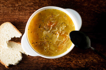 Chicken soup with noodles, potatoes, onions and carrots in a white deep plate with a black spoon and white bread on a wooden background.
