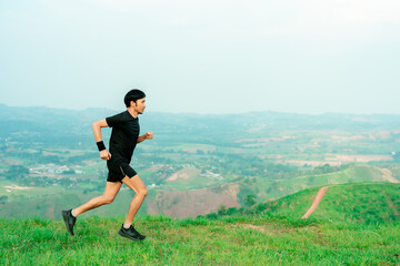 Fototapeta premium Young Asian man runner, wearing black sportswear, running on a big mountain trail, cool morning, windmills, and sky in the background.
