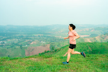 Fototapeta premium Young Asian woman runner, wearing black sportswear, running on a big mountain trail, cool morning, windmills, and sky in the background.