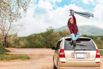 Fototapeta Happy teen girl is sitting on the roof of a car, joyfully raising her jacket. Country road at the background. Copy space. The concept of travel and obtaining a driver's license obraz