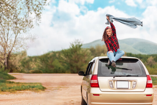 Happy teen girl is sitting on the roof of a car, joyfully raising her jacket. Country road at the background. Copy space. The concept of travel and obtaining a driver's license