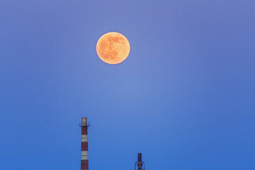 Fototapeta na wymiar A large moon in the evening sky above the industrial pipes of the plant.