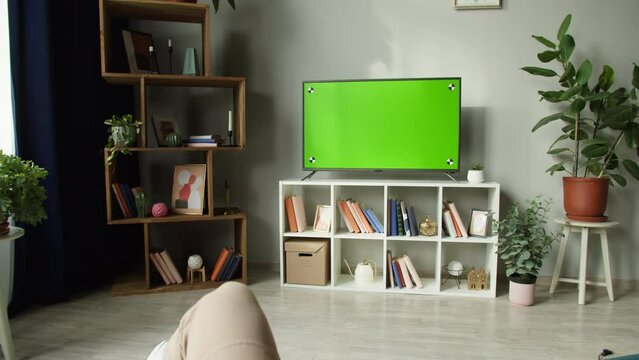 Woman watching television with chroma green screen. Unrecognizable female person switching channels on tv with remote controller, sitting on sofa in living room. Spare time at home, relaxing.