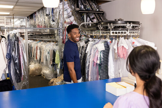Young man talking to customer at dry cleaners while he pulls clothes off rack.