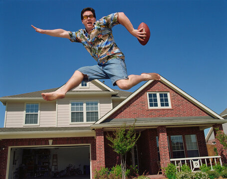Middle aged man holding a football jumping in front of home, striking a pose mid air