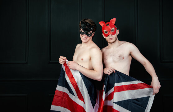 Portrait Of A Gay Couple In Red And Black Leather Masks Without A Shirt. Fashion Photo Of Men On A Black Background With The British Flag.