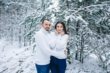 Fototapeta na wymiar winter love story. snow-covered pine forest. man and woman hugging in the winter forest. girl on a guy dressed in a white winter sweater in a white snowy forest
