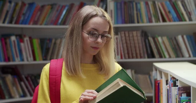 Rear of Caucasian young blonde beautiful woman in glasses walking in library passage, then choosing texbook from shelf and reading it. Female student flipping pages of book. Education concept.