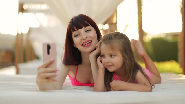Relaxed young woman and her daughter watching movie or having video chat on smartphone while lying in the resort gazebo near pool. Happy mother and her daughter sunbathing and holding mobile phone
