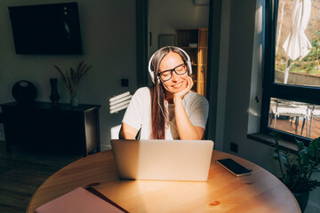 Young happy woman sitting at table at home in sunlight listening to audio in headphones in laptop.