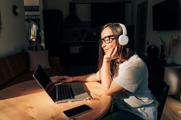 Woman sitting at a table at home in the sunlight listens to audio on headphones and uses a laptop.