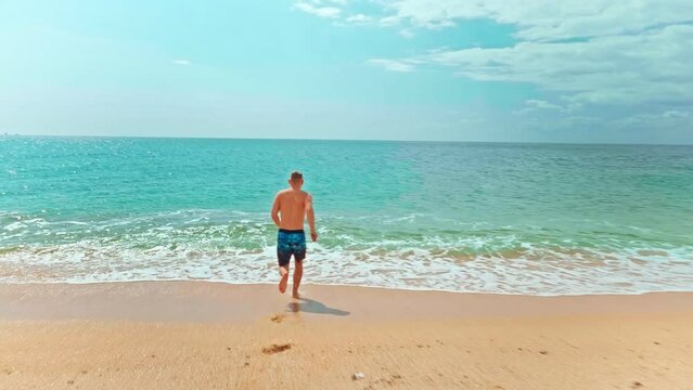 Slow-motion back view tracking shot of young man running along a sandy beach into the sea with clear blue water. Summer vacation on the sea concept. 