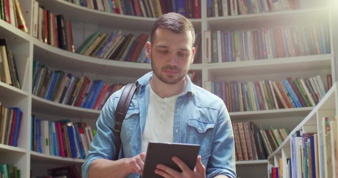 Caucasian young male student standing in library, using tablet device and tapping on screen. Stylish handsome man texting on computer and surfing in Internet online for information.