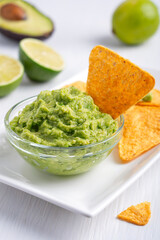 Guacamole Mexican homemade vegetarian avocado-based dip, spread, or salad made of mashed ripe avocado and lime citrus served in glass bowl on plate with tortilla chips as snack on white wooden table