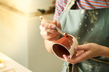 Close-up of girl painting clay mug with glaze. Woman coloring pottery in workshop with a paintbrush. Painter in green apron glazing clay pot. - 502635247