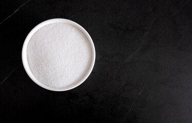 Granules of white sugar in a white bowl on a black background, free space