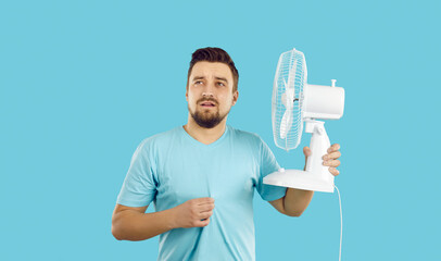 Fototapeta Man suffers from summer heat at home. Guy with broken air conditioner in his house using bad electric fan and sweating. Man in T shirt feeling hot and holding fan with sad face expression, studio shot obraz