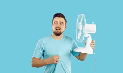 Foto op Plexiglas Man suffers from summer heat at home. Guy with broken air conditioner in his house using bad electric fan and sweating. Man in T shirt feeling hot and holding fan with sad face expression, studio shot © Studio Romantic