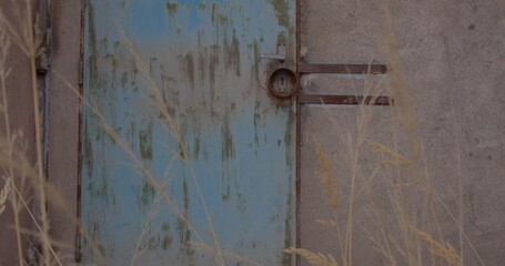 The lock on the metal door on the wall of the rear entrance of the building, the strips are attached to the wall. Concrete gray wall. Dry grass at the entrance to the facility. Scratches on the door.