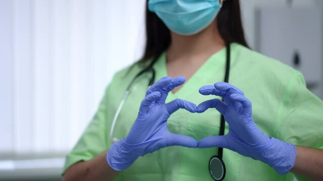 Unrecognizable female doctor gesturing heart shape at chest standing in hospital indoors. Young Caucasian woman in uniform coronavirus face mask and rubber gloves in medical clinic. Treatment concept