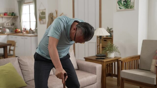 slow motion of Asian injured elderly female with back pain is walking slowly to the sofa with a crutch and sitting down for a rest.