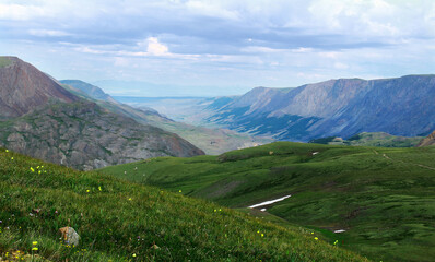 Panoramic distant view of a wide mountain valley with huge ridges, in the foreground green slopes with a road from the Karagem pass in Altai in summer, a sky with clouds