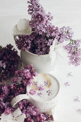 Obraz na płótnie Canvas Delicious coffee with lilac petals and vintage vase with blooming lilac branch on rustic white wooden background. Happy mothers day. Good morning. Beautiful lilac flowers and coffee still life