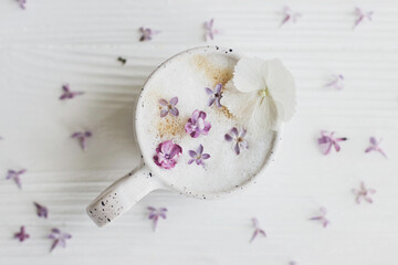 Delicious coffee with lilac petals on white wooden background flat lay. Happy mothers day. Good morning, welcoming spring. Beautiful flowers in stylish cup of coffee still life.