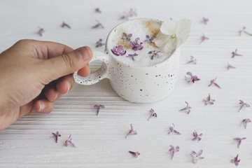 Hand holding delicious coffee with lilac petals on white wooden background. Happy mothers day. Good morning. Beautiful  flowers in stylish cup of coffee.
