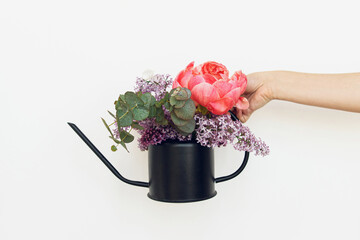 Hand holding stylish metal watering can with peony, lilac flowers and eucalyptus arrangement on...