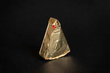 Triangular piece of melted cream cheese wrapped in gold aluminum foil on black background....